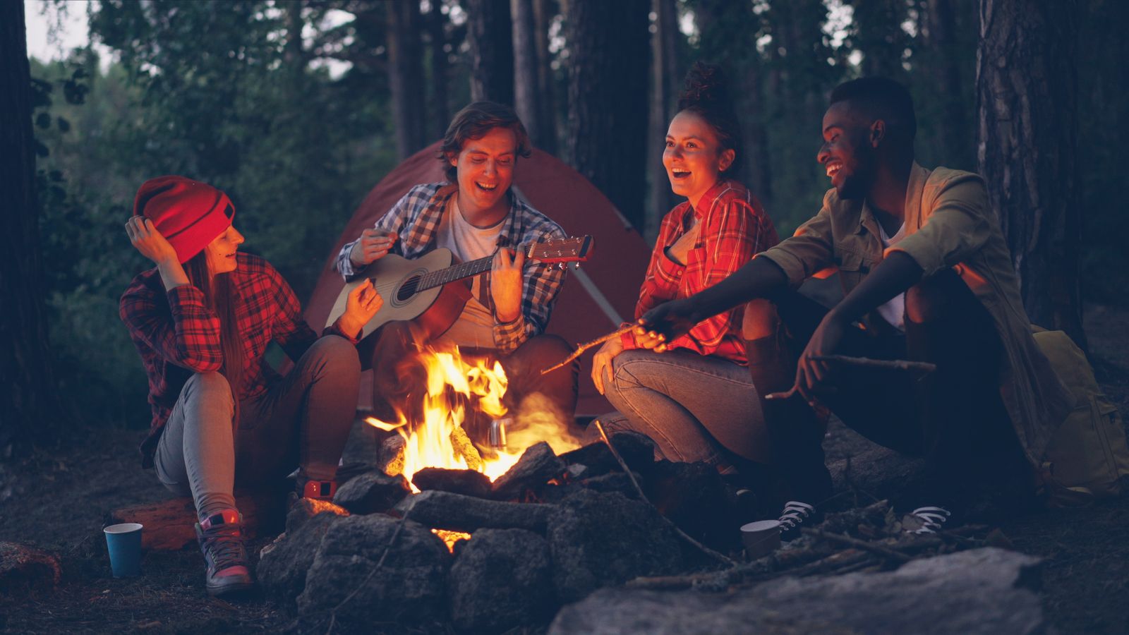 Un grupo de amigos alrededor de un fogón con una guitarra, junto a una carpa en el bosque
